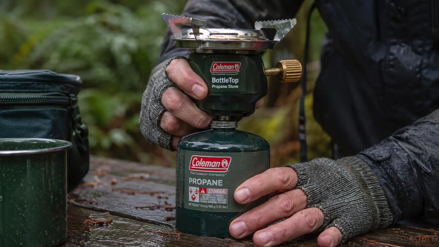 Close-up of a gloved hand threading a green Coleman BottleTop stove onto the top of a propane canister, the burner ring and adjustable knob in focus.